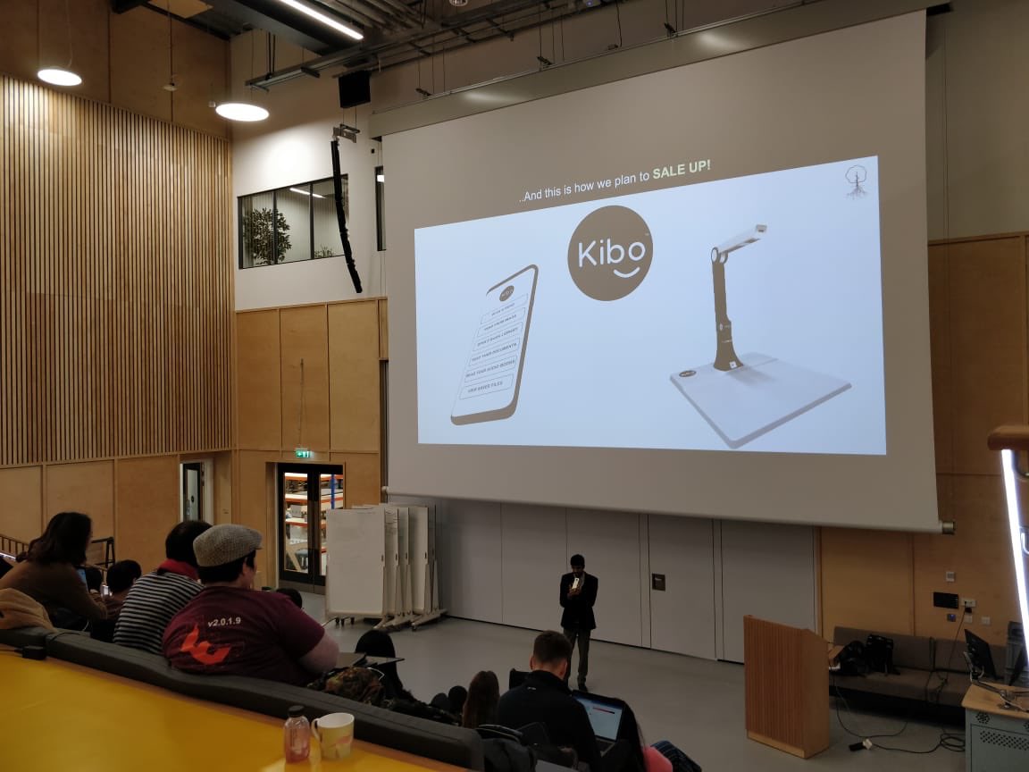 Man presents to an audience in a lecture theatre. Screen shows a picture of an assistive technology device.