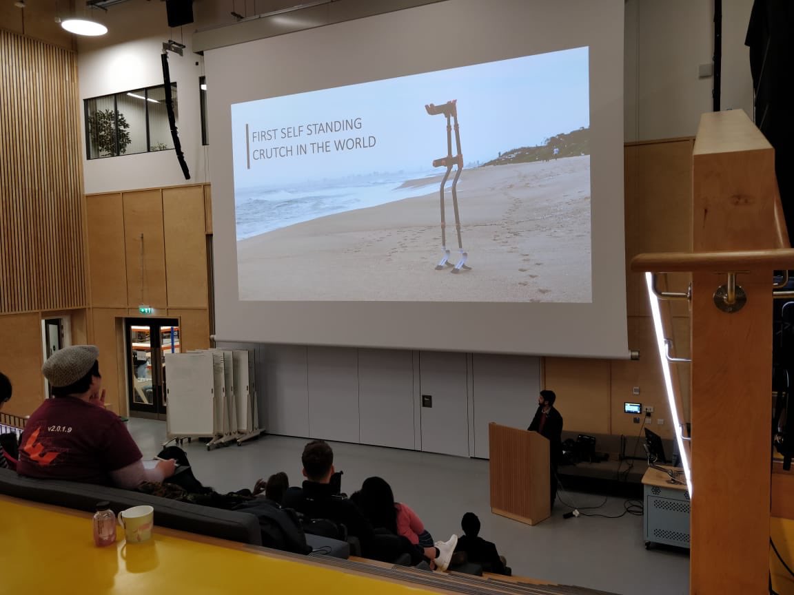 Picture of lecture theatre. Man stands in the podium, the audience is in the background. The screen shows a picture of a standing wheelchair.