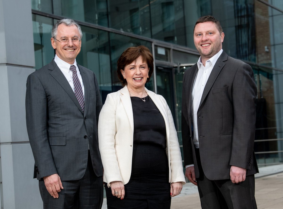 Two men and one woman standing are smiling in front  of a glass building.