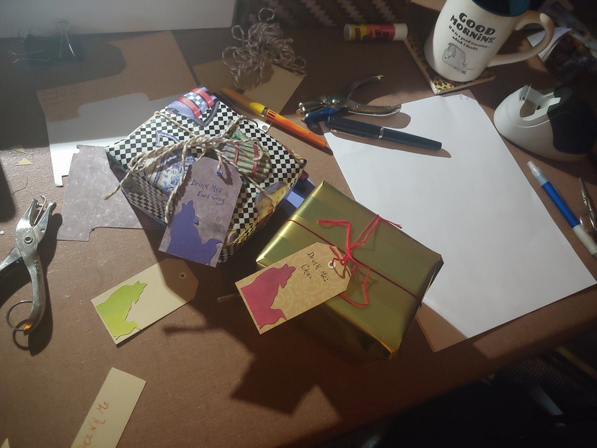 A card table covered in crafting detritus and fountain pens.  Two wrapped packages in the center of the chaos labeled "drink me: earl grey" and "drink me: chai"