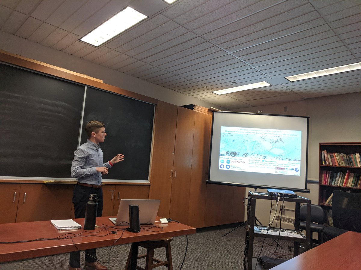 Photo of a person standing in front of a blackboard in a classroom and gesturing towards a projected slide with a title and glacial image.