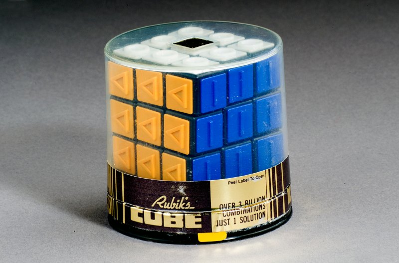 The image shows the yellow and blue faces of a Rubiks cube still in its original packaging, although the seal has been broken.