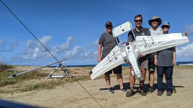 Four scientists hold a winged drone on the beach. 