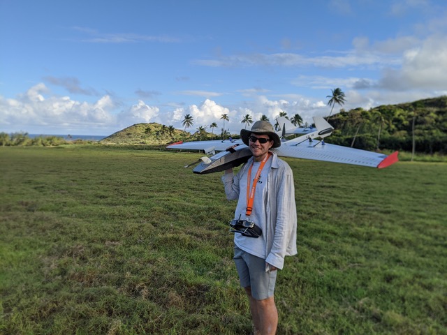 Man holds a winged drone (shaped like a small glider plane) over his shoulder, with grass and palm trees in the distance