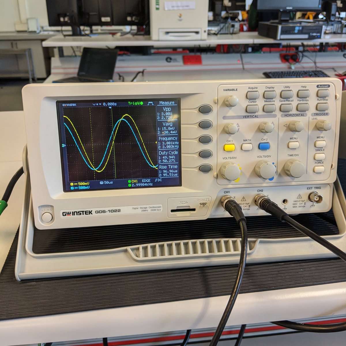 A white oscilloscope with grey, yellow And blue buttons...some dials. This time, the blue output signal is only slightly smaller in amplitude when compared to the yellow input signal. The blue wave lags behind the yellow wave, but not as much.