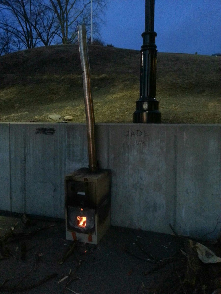 a nighttime photograph of a filing cabinet that has been turned into a (lit) furnace with a tall chimney. the furnace is next to a concrete wall, on which jade's name has been written in charcoal