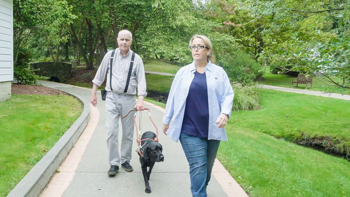 Curt & black lab guide dog Quincy walk on the nature path with Mobility Instructor, Woody. Woody demonstrates how walking ahead of the team may cause confusion.