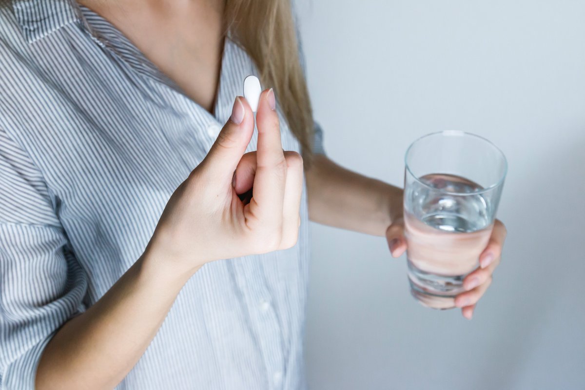 A woman holds a white pill in one hand and a glass of water in the other