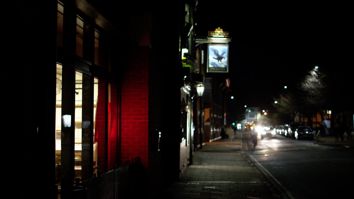 A brick wall is lit up by a red sign; in the background: a pub sign, and oncoming traffic