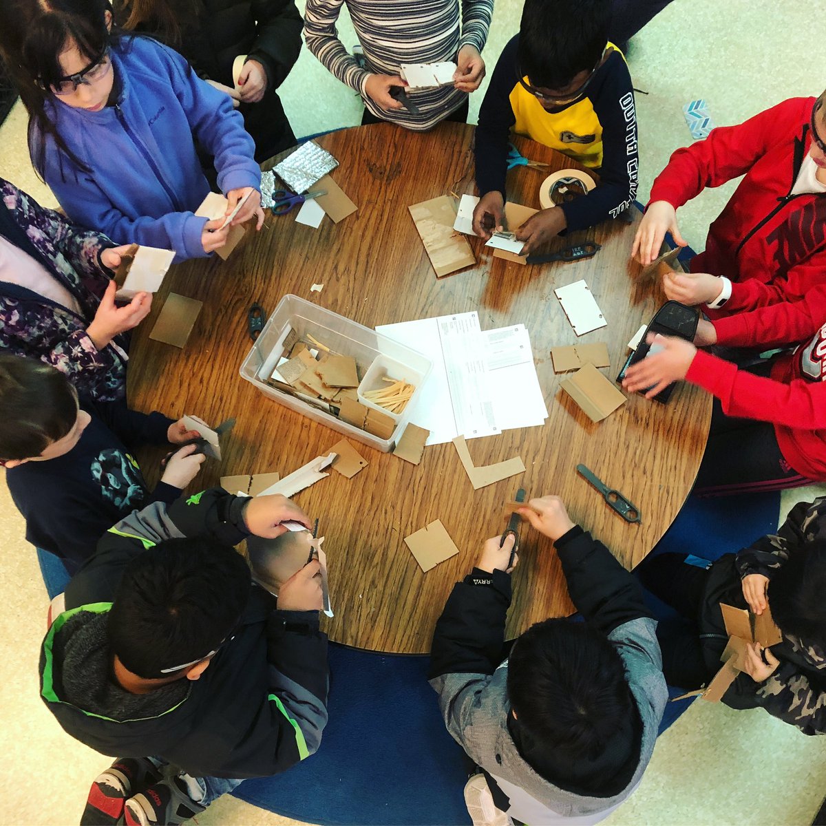 11 students working at a round table, cutting and building with cardboard.