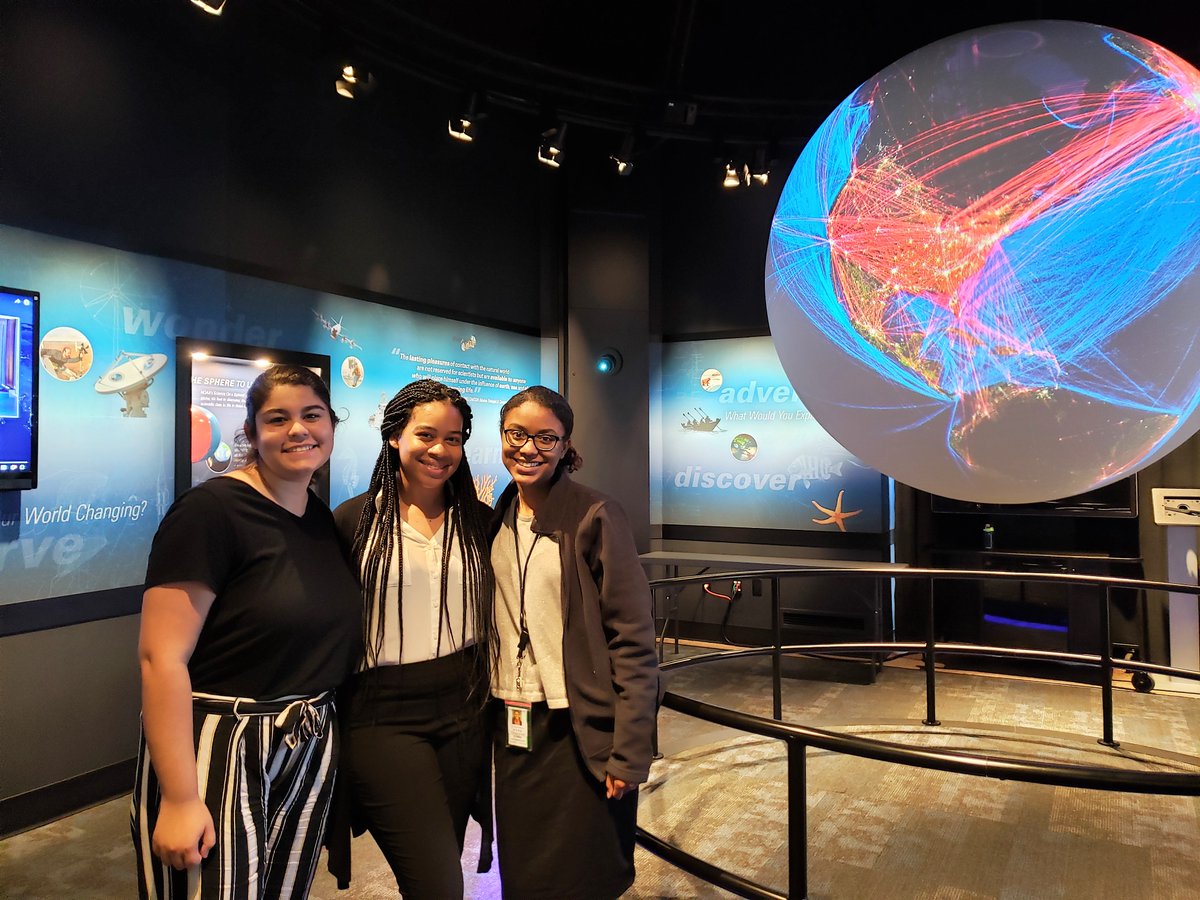 Three women stand next to a large sphere which is displaying human transportation data.