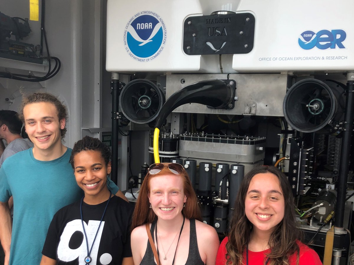 Four students stand in front of a large piece of machinery which has a NOAA logo on the front.