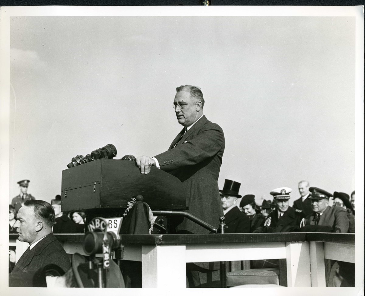 Photo of President Roosevelt standing behind a podium giving a speech with dignitaries sitting in the background.