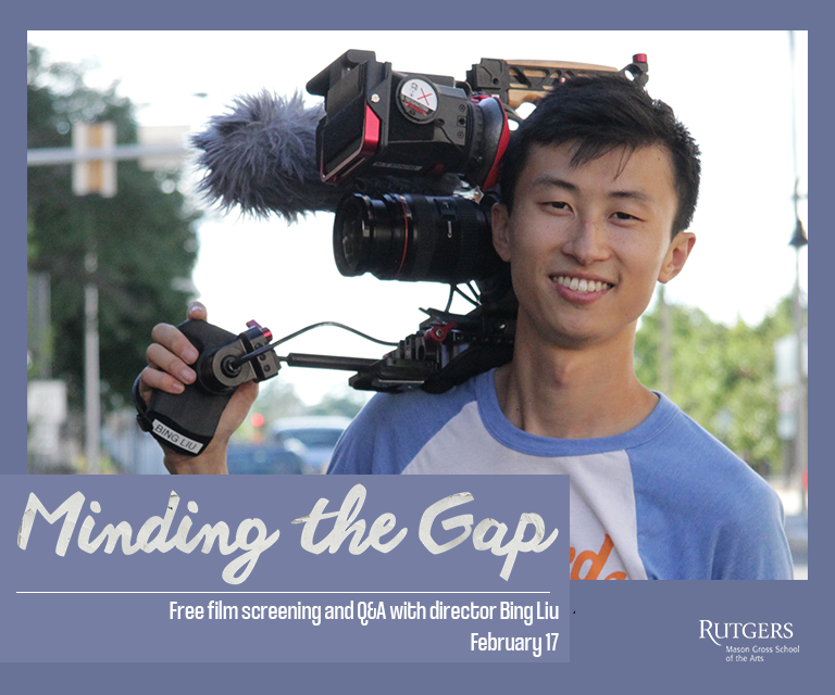 Young man with a video camera on his right shoulder, smiling, in a T-shirt with trees and a city street behind him.