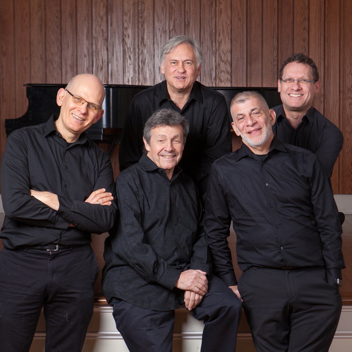 Five men in late middle page, dressed formally, grouped together at the lip of a stage in front of a piano, smiling. 