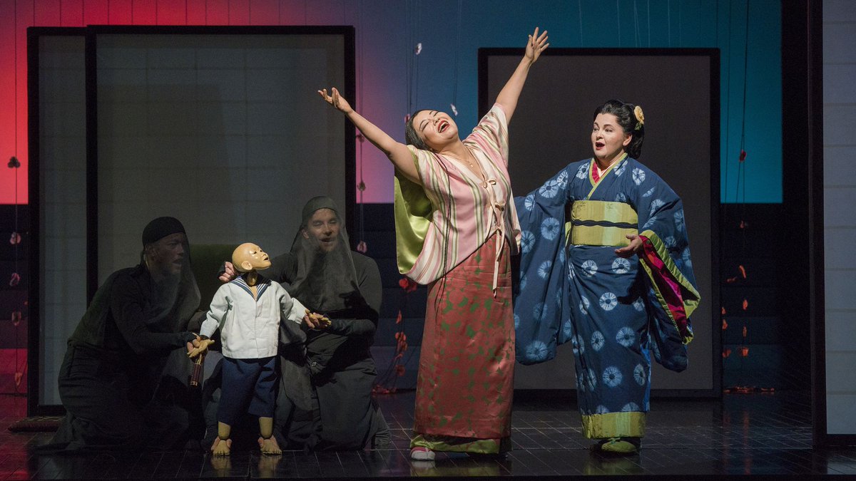 A woman extends her arms upwards joyously, while a second woman comes to embrace her and a young boy puppet manned by two actors looks on.