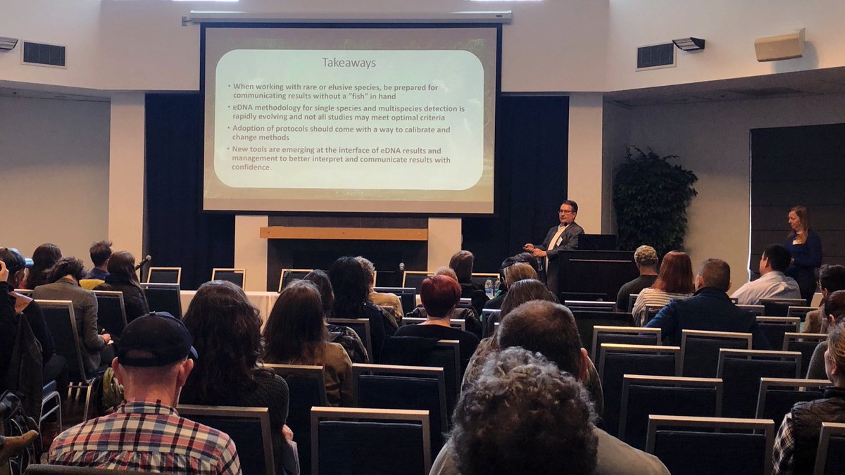 An audience looks at a man speaking at the front of a room.