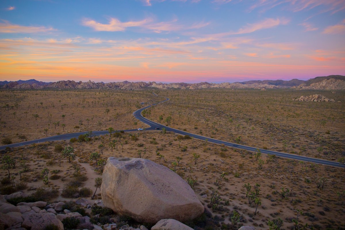 The sun sets over a view of a faraway road, Joshua trees, and granite boulders.

Photo: NPS / Glauco Puig-Santana