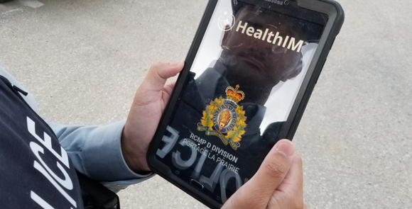 A male RCMP officer holding a tablet outdoors with his reflection visible on the screen.