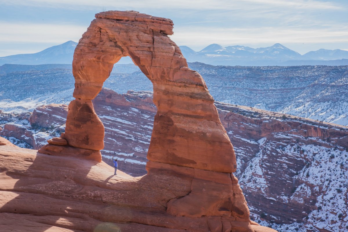 A tall red rock arch stands on a plateau with snow covered mountains in the background.