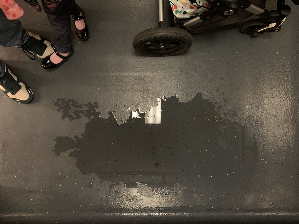 A puddle of urine pools in a BART elevator next to a stroller. Also visible are the shoes of an adult and a 5-year old.