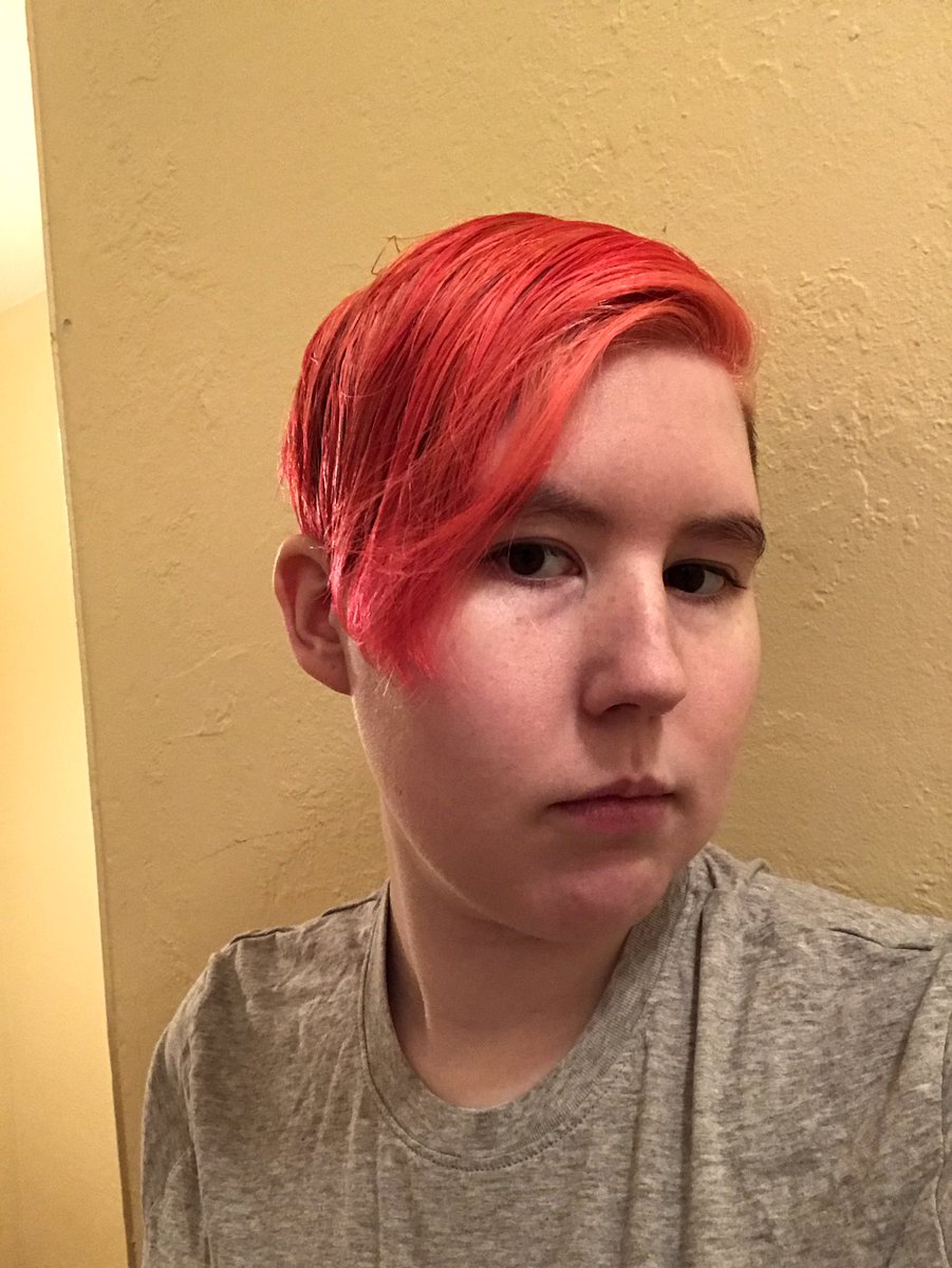 A picture of a white person with pink hair. They are wearing a grey shirt and standing in front of a yellow wall.