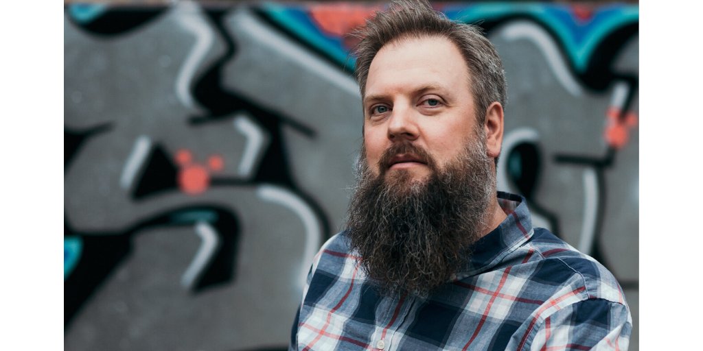 Photo of Jason Dorwart, a white male about 40 years old with a long beard and greying hair looks at the camera. He wears a blue and red plaid shirt. Behind him is a mural silver, pink, and blue graffiti lettering.