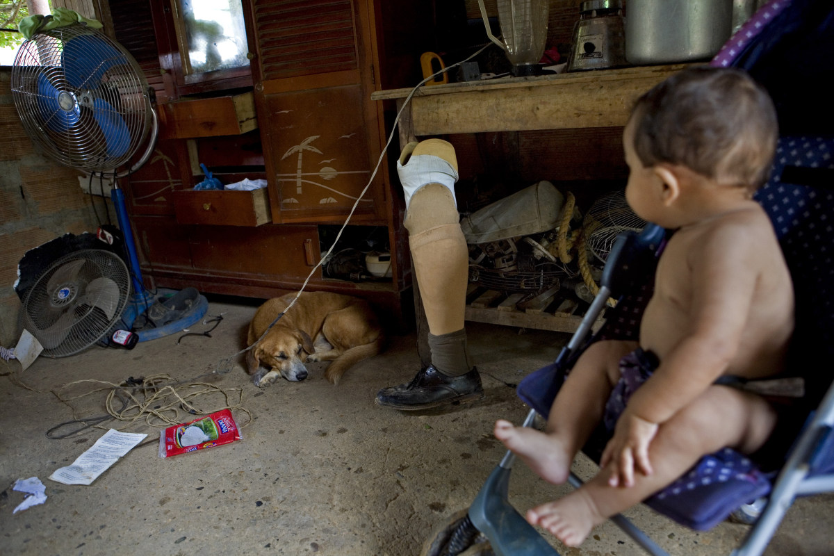 A baby in Colombia sits in a pushchair, and looks over his right shoulder toward a dog on a concrete floor, as well as his father's artificial leg, which rests against a cabinet. There is a broken fan on the ground, as well. 