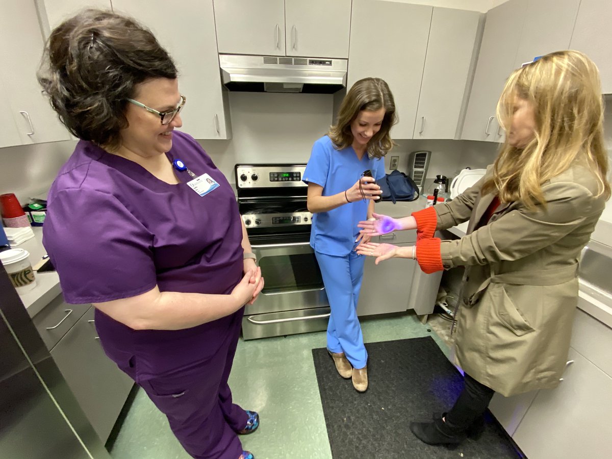 A employee holds out her hands under a black light for a hand washing demonstration as two nurses look on.