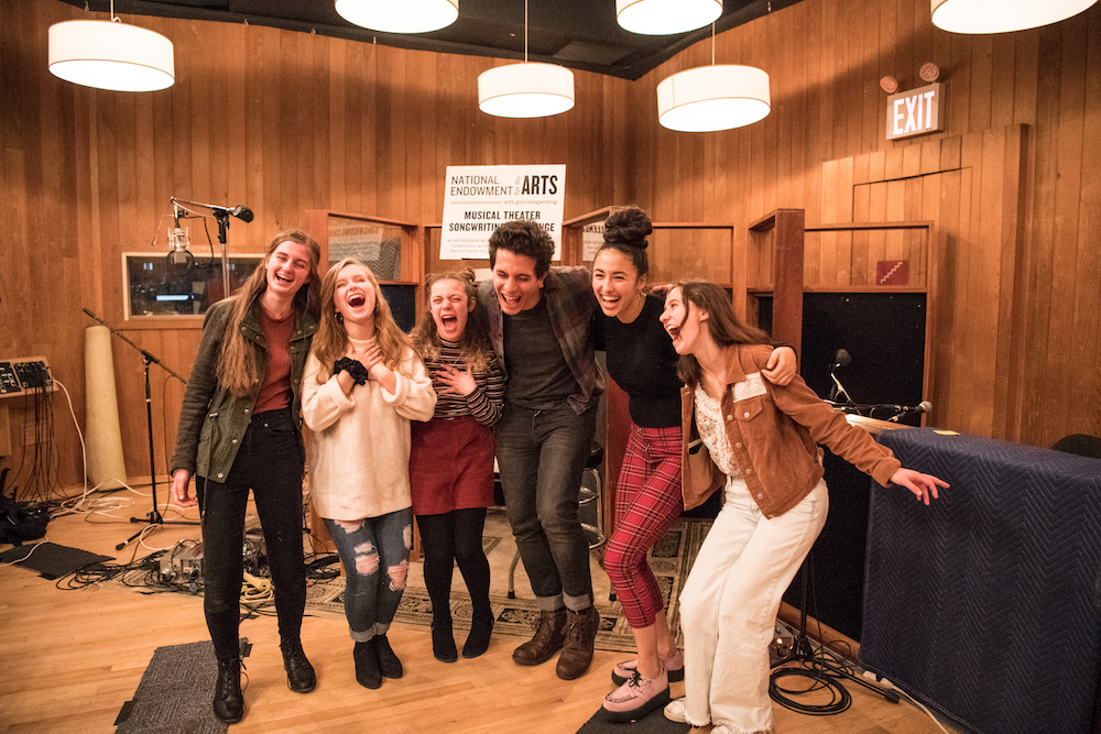 A group of teens stand next to each other with their arms around each other laughing as they pose for a photo