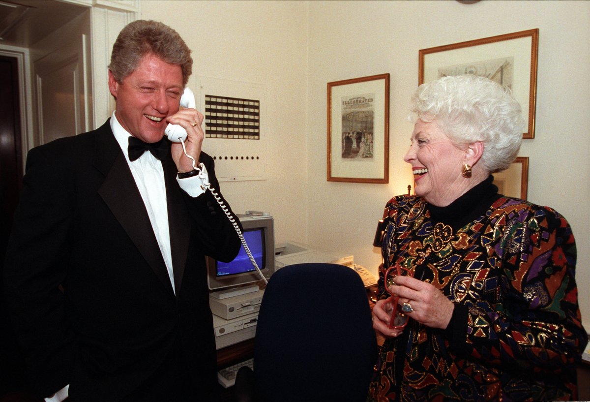 President Clinton, dressed in a tux, laughs as he speaks into a white corded telephone. Texas Governor Ann Richards also laughs as she stands nearby.