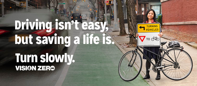 Woman holding up a sign informing driver's to turn slowly and yield to cyclist. Text reads: Driving isn't easy, but saving a life is. Turn Slowly. VISION ZERO