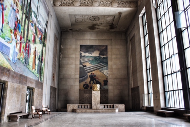 A view of the Shrine of the Immortal memorial hall at the Worcester Memorial Auditorium. The mural opposite the viewer depicts American sailors on a #Navy ship in a stormy sea. At left is a partial view of another mural, depicting a young woman, surrounded by other people, laying a wreath on the grave of a fallen soldier. At right are floor-to-ceiling windows. 