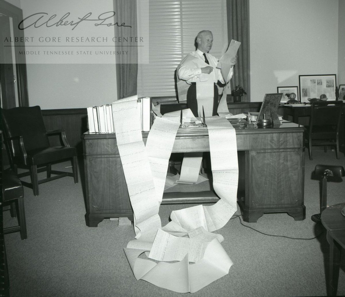 Black and white photo of Senator Gore standing behind his desk. Draped over his desk and around him is one long connected sheet of white paper.