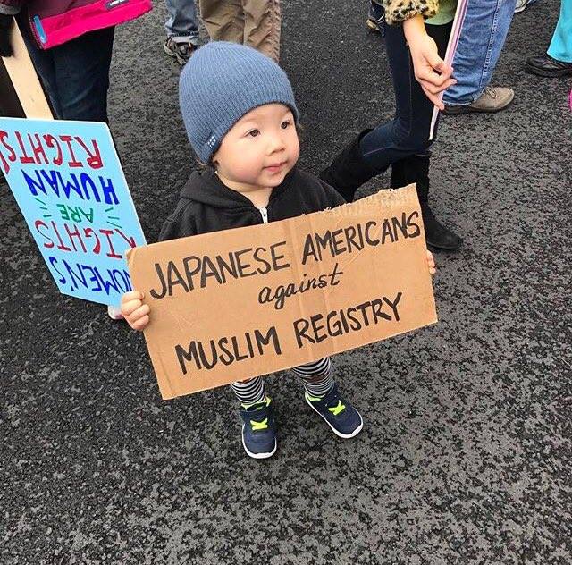 2 year old child holding a cardboard sign at a protest that reads "Japanese Americans against Muslim registry"