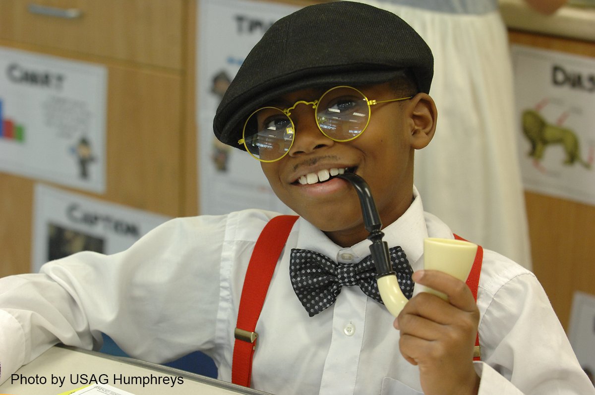 A youg boy is dressed up as a 100-year-old man, wearing a bowtie and hat, glasses and holding a pipe in his mouth.
