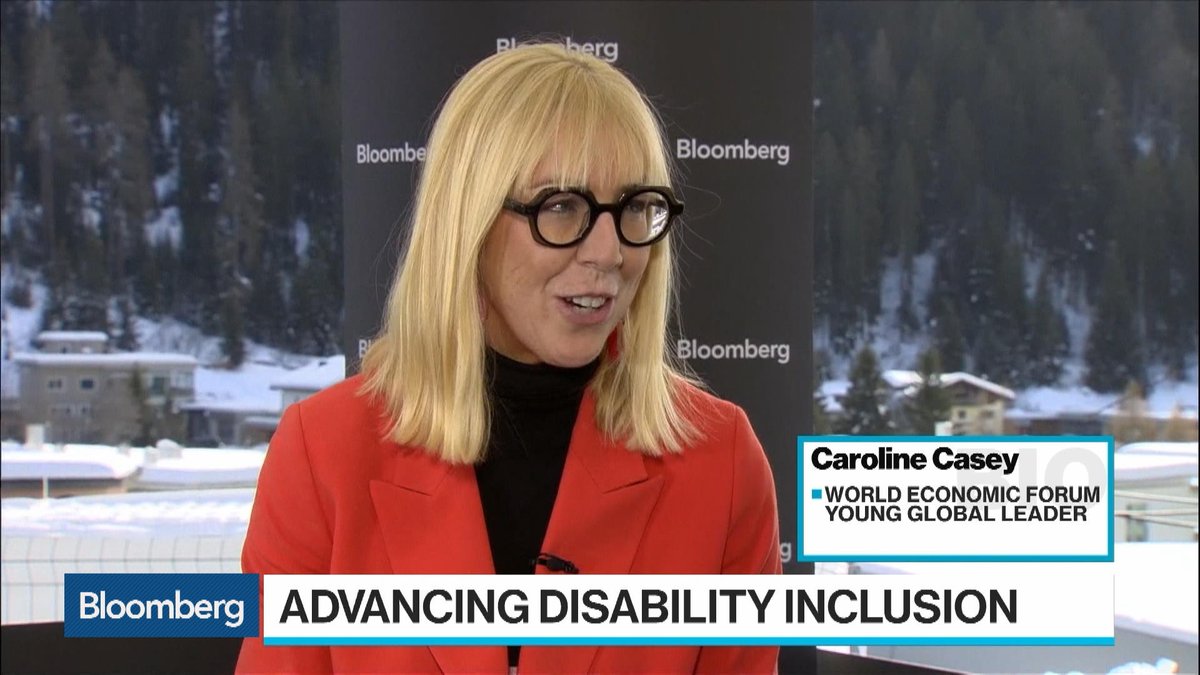 Seated woman wearing a red suit jacket, blonde hair with front fringe and black circle glasses. Background is a snowy mountain with trees and Bloomberg signage. Bloomberg: Advancing Disability Inclusion. Caroline Casey, World Economic Forum, Young Global Leader. 