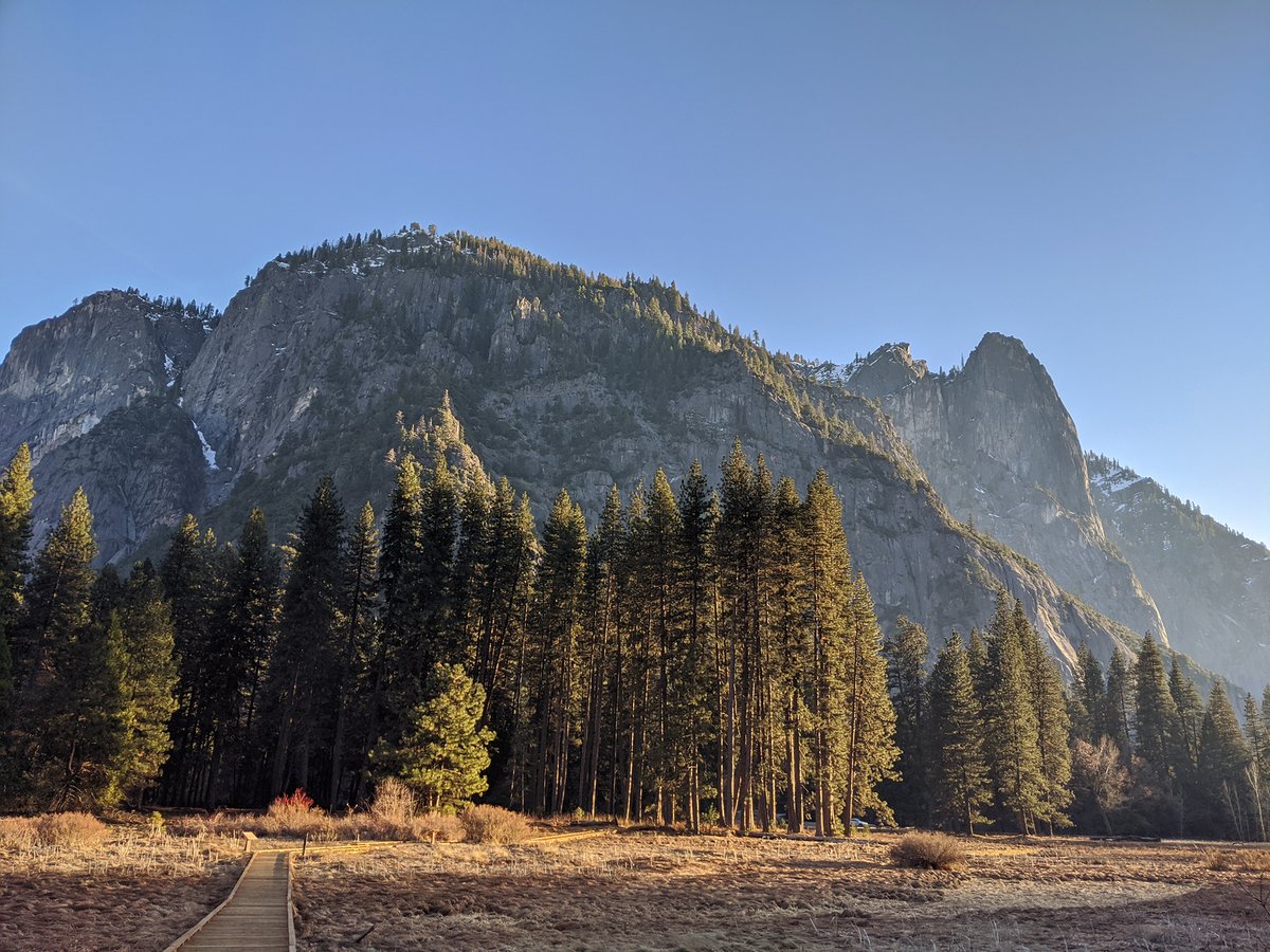 A wooden boardwalk crosses the meadow to a cluster of trees. Granite formations rise in the background.