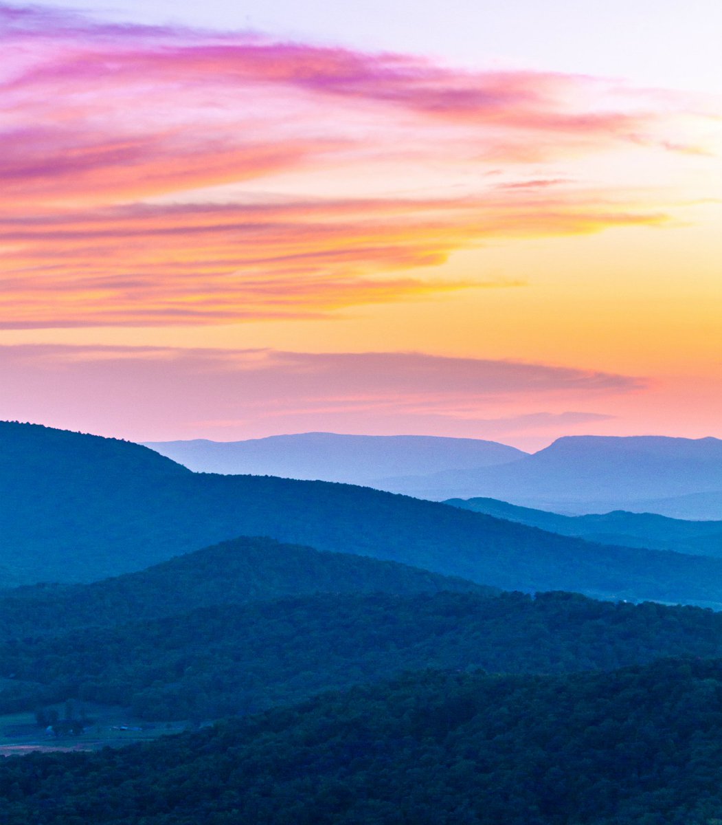 A series of low mountains run back to the horizon under a colorful sunset sky.