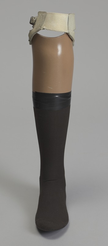 A solid fiberglass prosthetic left leg (including below knee, calf, ankle), and foot. The leg, or laminated frame, is a light brown, with a calf-high black sock taped up around the calf with black tape. The top of the leg, or frame, is cut to fit around the knee (cut lower in the back for joint movement and bending at the knee) and has a leather harness attached, creating a flexible inner socket-like formation. The