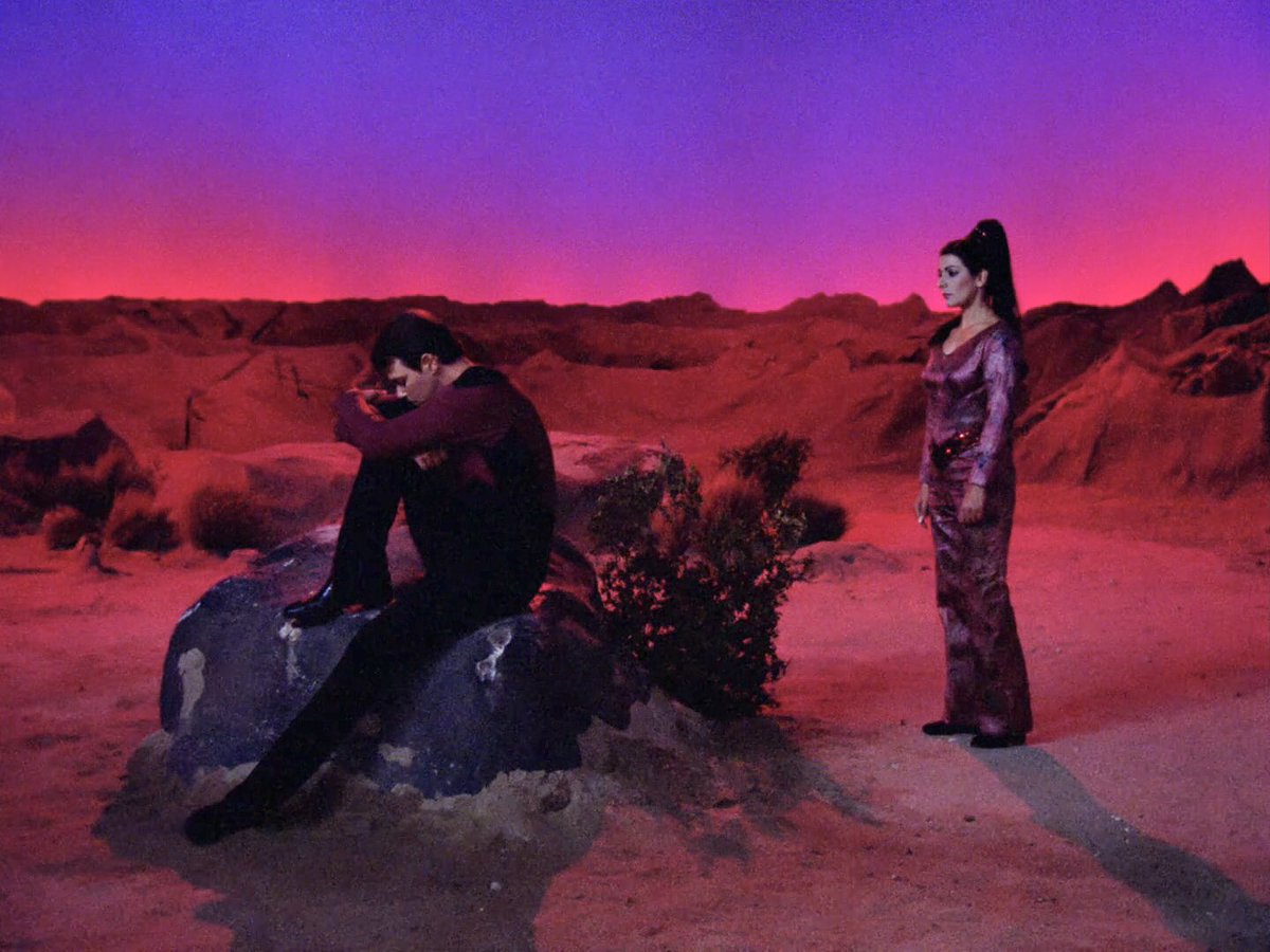 Riker looking sad and pensive on a rock in a colorful, sunsetty desert setting with Deanna Troi nearby