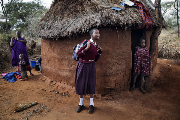 Image shows a girl wearing her school uniform as she puts on her backpack. She's outside her family's hut.