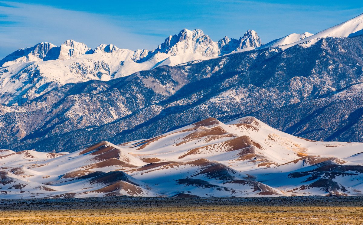 Tall sand dunes are dusted with snow as a large snow covered mountain range dominates the horizon.