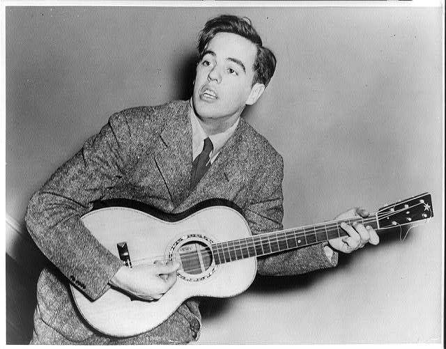 A young Alan Lomax playing acoustic guitar. Black and white image. It's a candid shot. He's in mid strum and mid song.