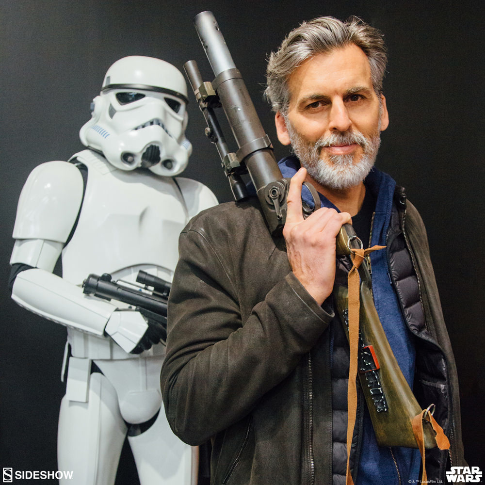 Sexy salt and pepper AND BEARDED  Oded Fehr wears rugged earth toned layers and heavy leather jacket while holding a Star Wars black market blaster a storm trooper life size figure is behind him