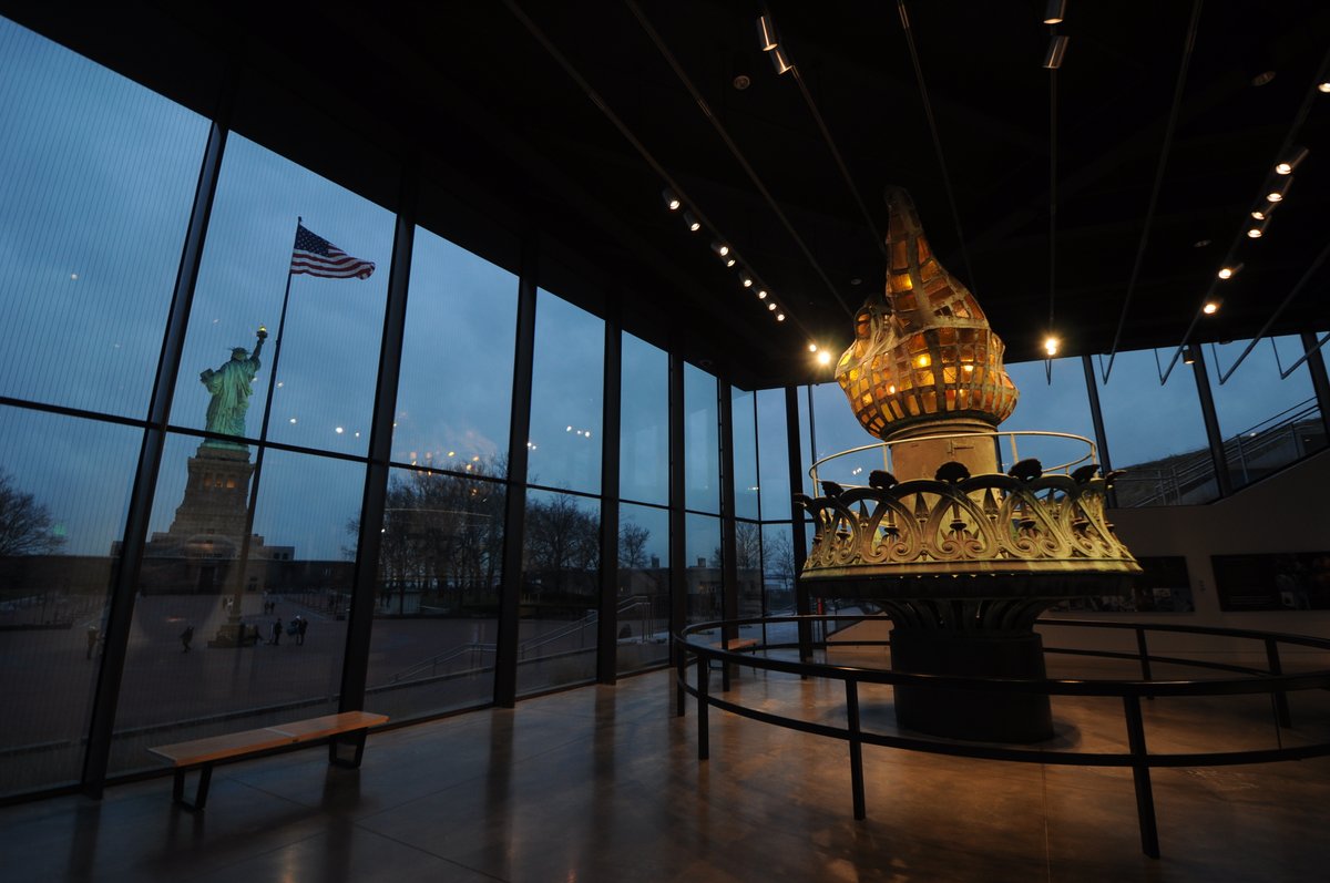 The original torch in the museum is illuminated while the sun goes down on the Statue of Liberty in the background.