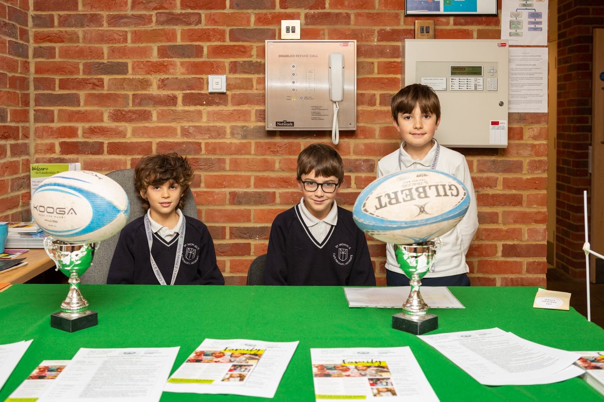 Three boys sit at a desk decorated with rugby balls and trophies.