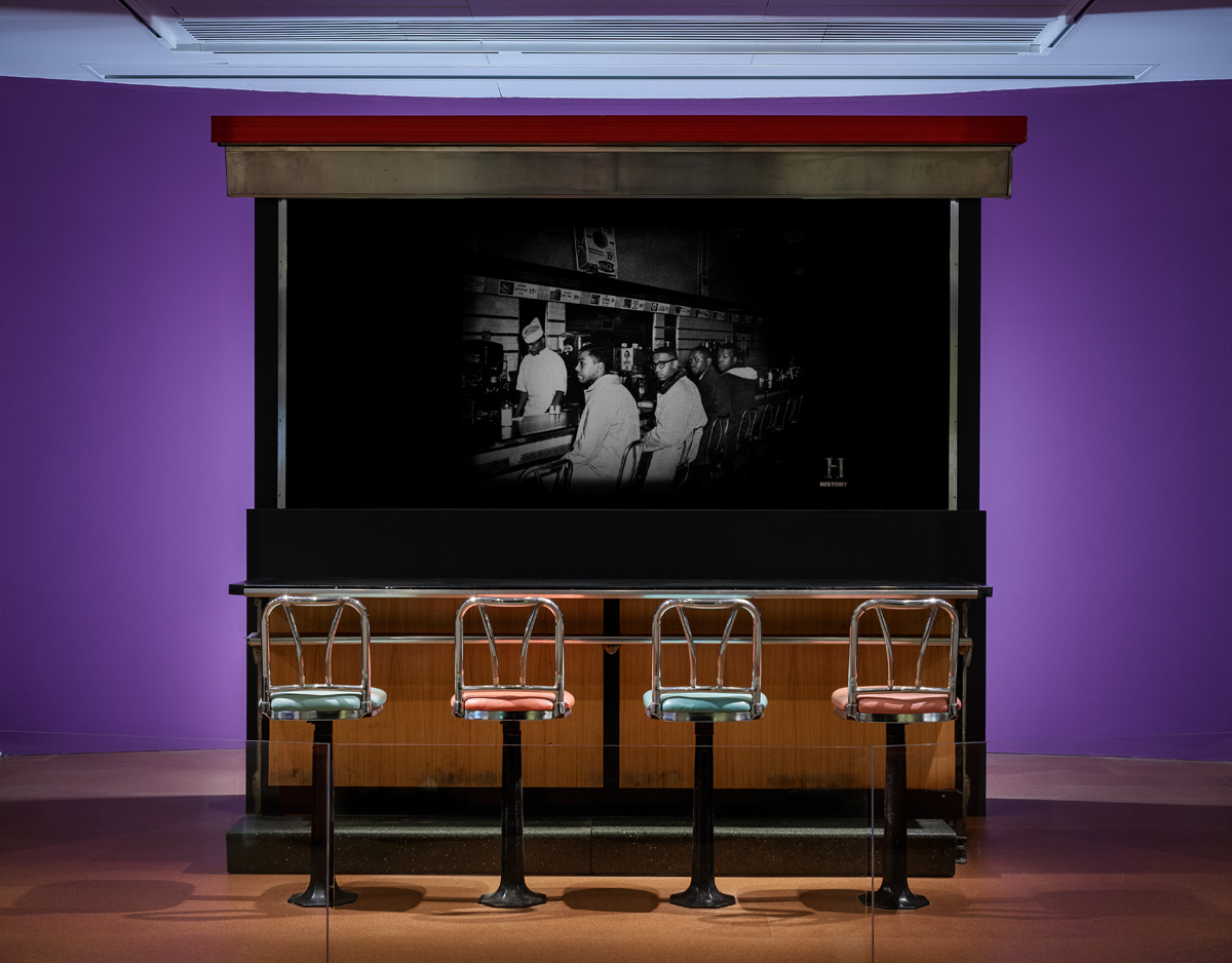 Counter with four stools in front of it. Behind the counter is a black and white image of two young men sitting at a lunch counter.