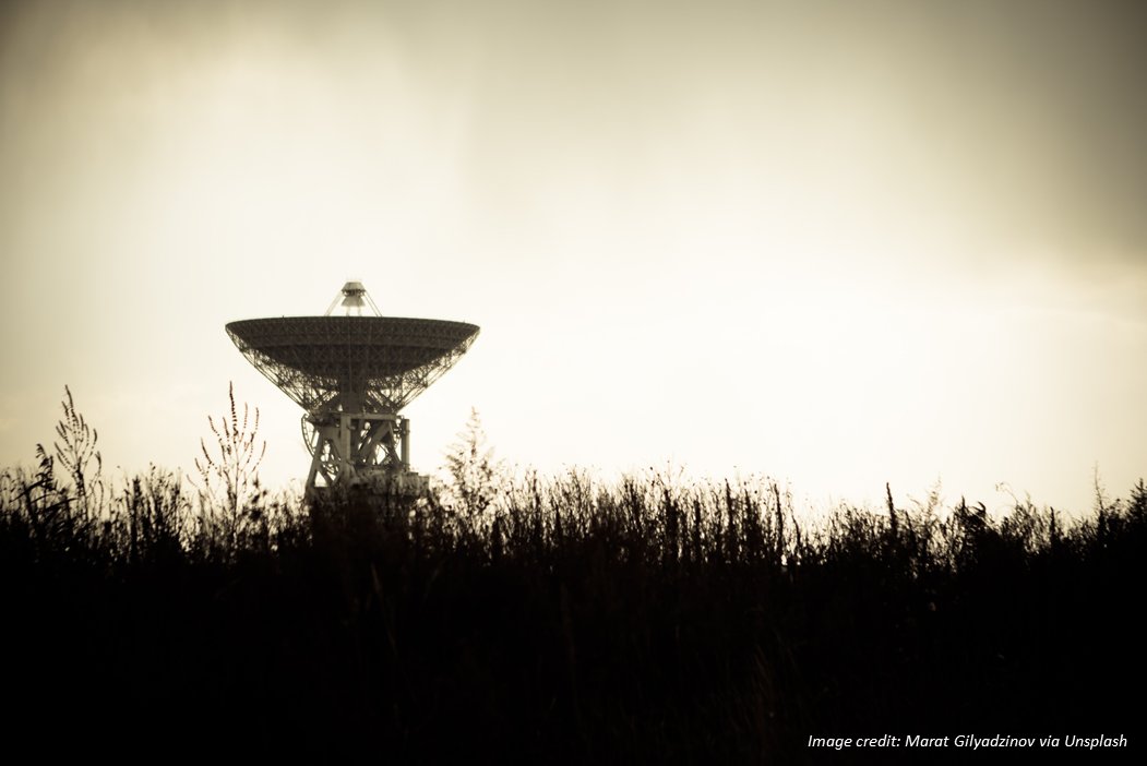 Silhouette of radio telescope dish against lighted horizon seen from across an open field. Image credit: Marat Gilyadzinov via Unsplash.