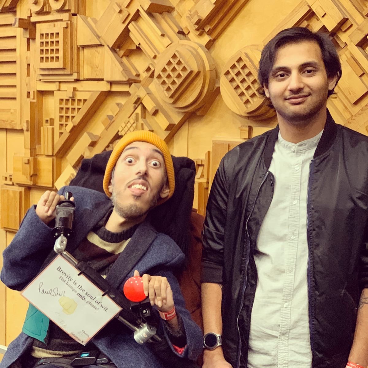 Sulaiman (who is sitting in his power wheelchair and he’s wearing his orange beanie hat, he dark blue wool blazer, his brown jumper with stripes, and his navy chinos) is making a silly face with big eyes next his lifts brother Sufian (who a South Asian man with black hair and he’s wearing a black casual jacket, a white shirt, and light blue jeans). Both are very happy.